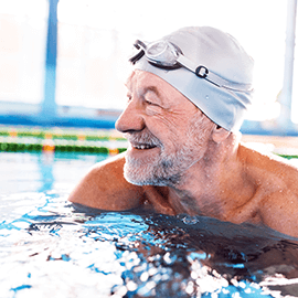 Smiling older man in a swimming pool wearing a grey swim cap and goggles